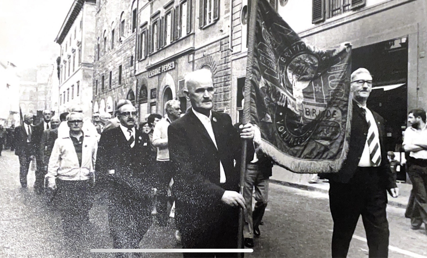 Charlie Hutchison (background, white shirt, side profile) walking in a political demonstration. Foreground shows an International Brigade banner. Unknown date and location.